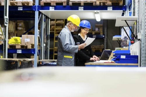 Man and woman both in hard hats in a warehouse reviewing paperwork
